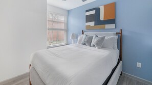 Ein Schlafzimmer mit Bett, einem Fenster mit Jalousien, einem an der Wand befestigten abstrakten Kunstwerk und einem Nachttisch mit Lampe.