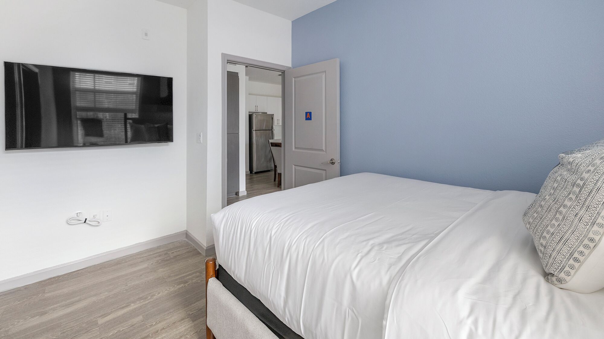 A bedroom with a bed, a TV mounted on the wall, and a door leading to a kitchen.