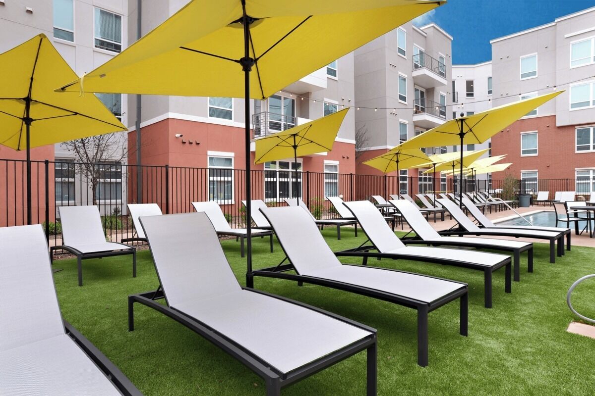 Cadeiras de descanso ao ar livre com guarda-sóis amarelos, uma piscina e um edifício com varandas ao fundo.