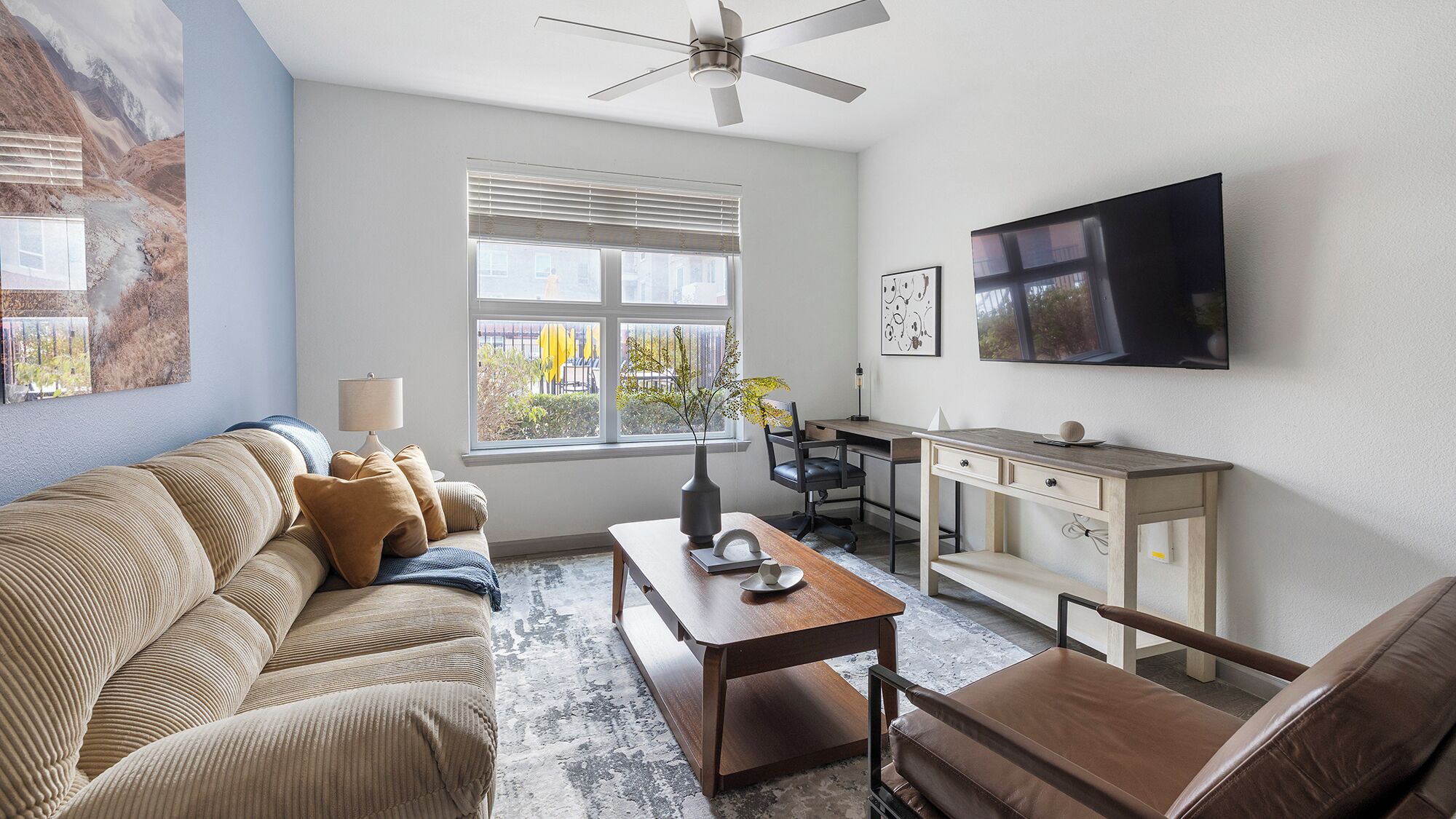 A living room with a sofa, coffee table, and a desk with a chair. There is a window with blinds, a ceiling fan, and a flat-screen TV mounted on the wall.