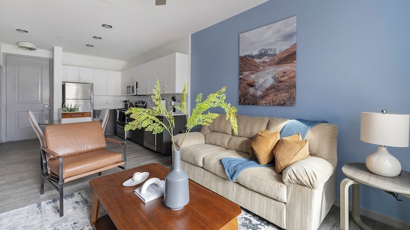 A modern living room with a leather chair, a sofa with cushions, a coffee table, and a vase with a plant. There is a kitchen in the background with white cabinets and a microwave. - Landing Apartments Downtown Arlington (Arlington)
