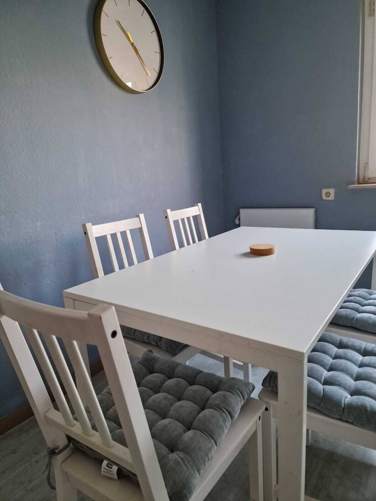 A dining area with a white table, four white chairs, a clock on the wall, and a window with curtains.