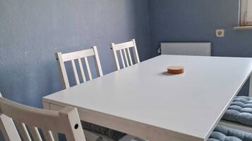 A dining area with a white table, four white chairs, a clock on the wall, and a window with curtains.