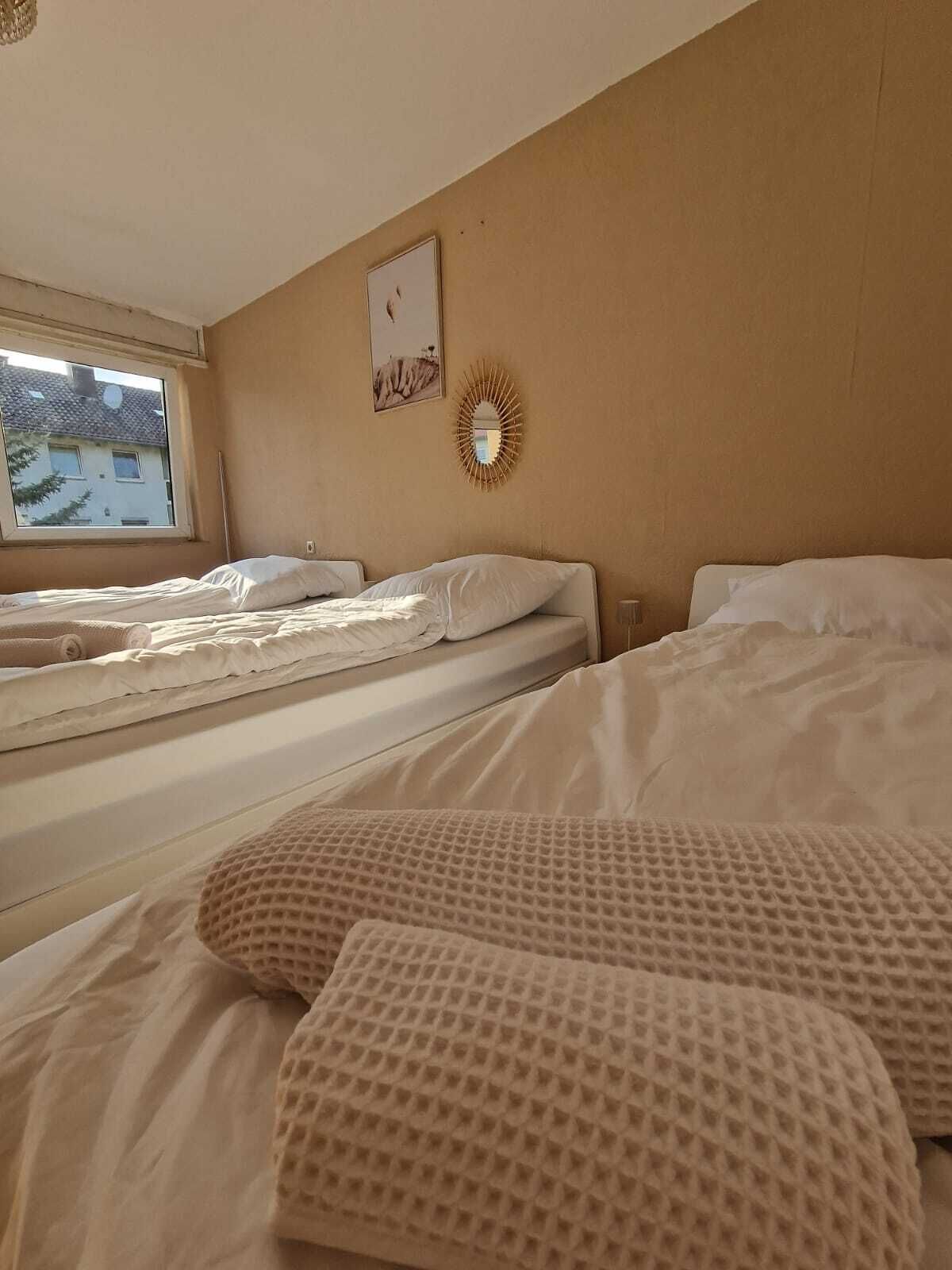 A room with two beds, a window with a view of buildings, a wall-mounted mirror, and a wall clock.
