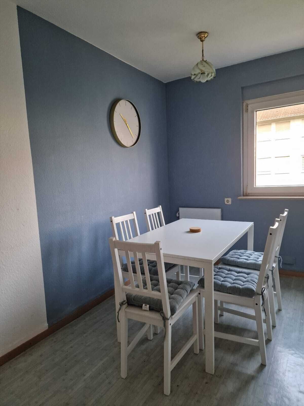 A dining area with a white table and chairs, a clock on the wall, and a window with blinds.