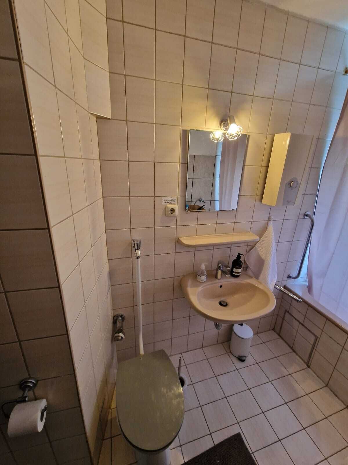 Un baño pequeño con inodoro, lavamanos y espejo. Hay un perchero sobre el lavamanos y una ventana con cortina.