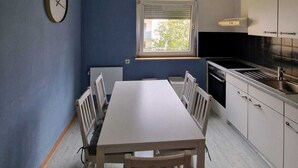 Eine kompakte Küche mit Essbereich, ausgestattet mit einem weißen Tisch und Stühlen, einer Wanduhr und einem Fenster mit Jalousien.