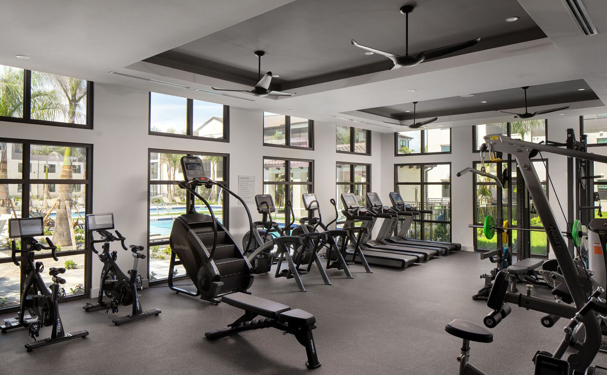 Ein Fitnessstudio mit verschiedenen Trainingsgeräten, großen Fenstern und Blick auf ein Schwimmbad und Palmen.
