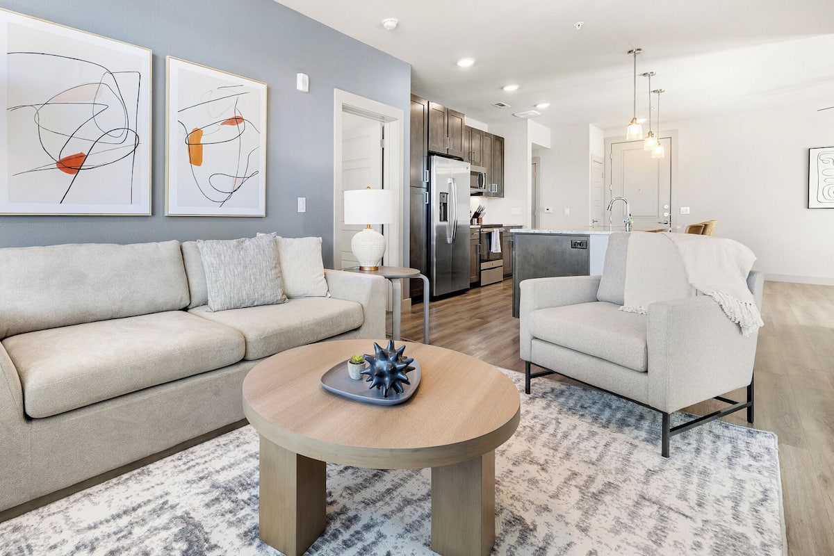 A modern living room with a sofa, armchair, and coffee table, a kitchen in the background, and abstract art on the walls.