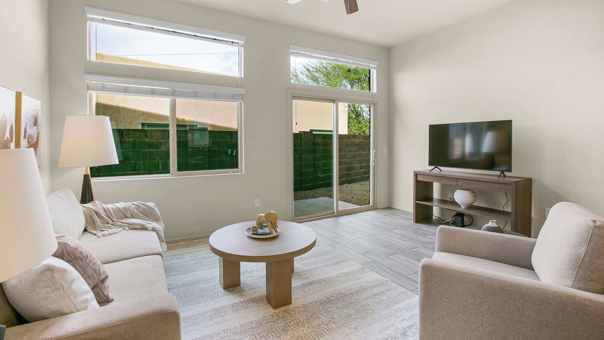 A modern living room with a sofa, armchairs, a coffee table, a TV on a wooden stand, and large windows with a view of a green area.