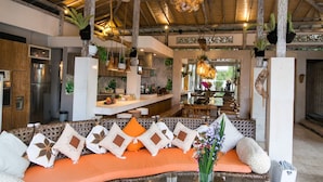 Een ruime woonkamer met een houten plafond, een glazen salontafel en een zithoek met oranje kussens.