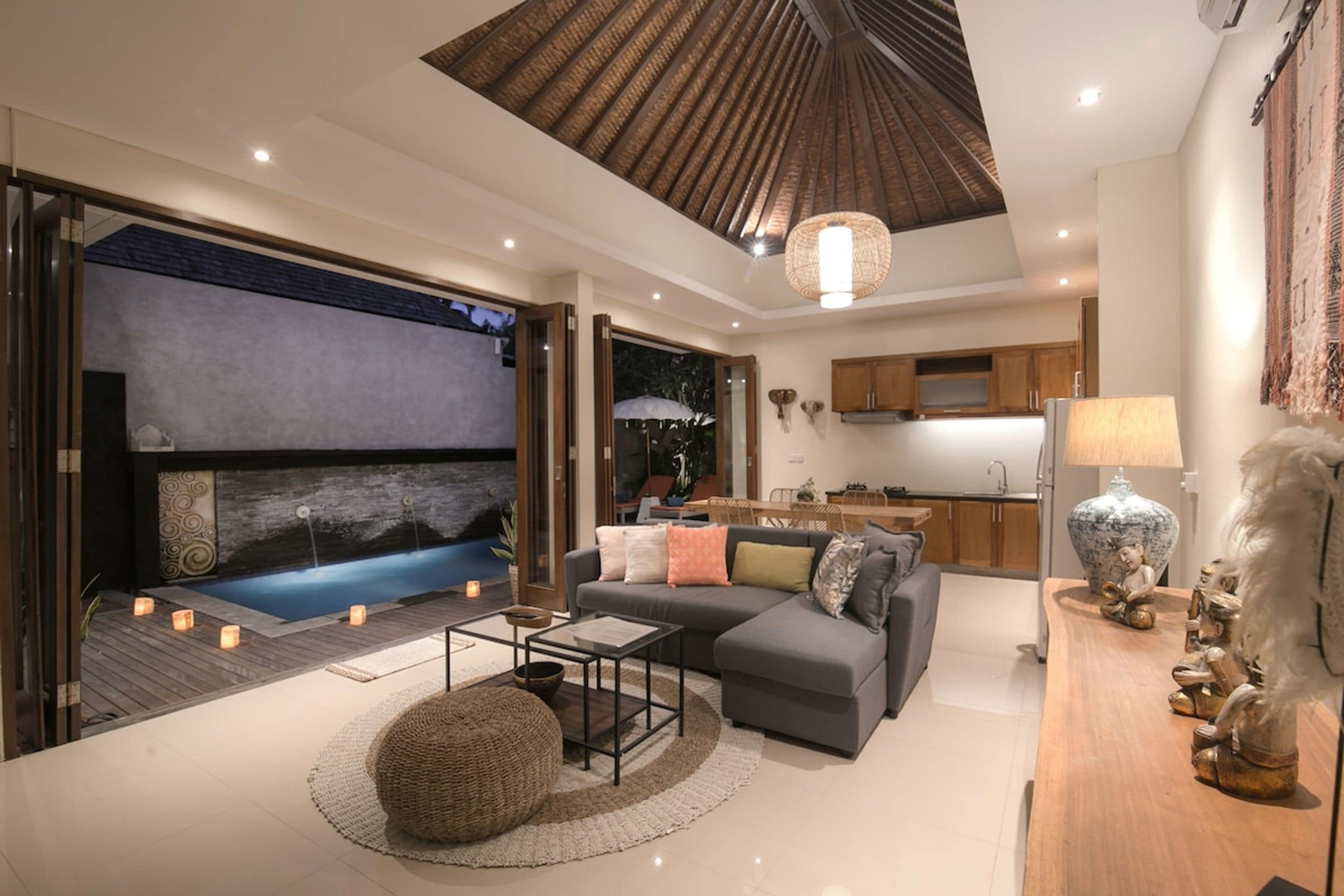 A modern living room with a grey sofa, a wooden coffee table, and a round pouf. There is a kitchen area with wooden cabinets and a dining table. A large window provides a view of the outdoors.