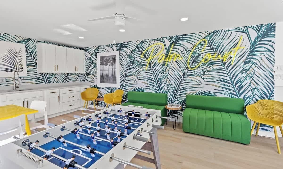 A modern living room with a foosball table, green sofa, yellow chairs, and a wall with a tropical leaf pattern.