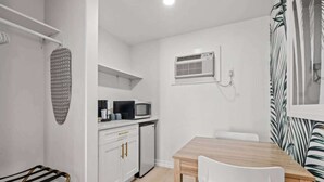 A compact kitchen with white cabinets, a microwave, and a small refrigerator. There is a wooden dining table with two chairs and a hanging drying rack. - Sunny Studio Near the Beach at Palm Court (#12) (North Myrtle Beach)