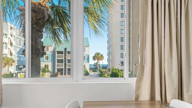 Una habitación de hotel con un ventanal grande, una mesa de madera, una silla blanca y cortinas color beige.