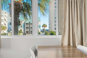 A hotel room with a large window, a wooden table, a white chair, and beige curtains. - Sunny Studio Near the Beach at Palm Court (#8) (North Myrtle Beach)