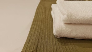 A neatly made bed with a green bedspread and white towels, a white headboard, and a window with curtains.