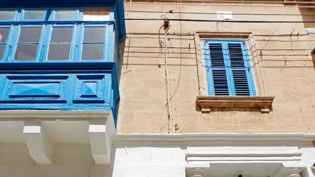 A building facade with a white wall and blue shutters, a central arched doorway, and a small window above the door.