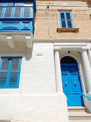 A building facade with a white wall and blue shutters, a central arched doorway, and a small window above the door.
