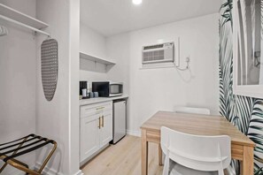 Una cocina compacta con armarios blancos, un microondas y un refrigerador pequeño. Hay una mesa de comedor de madera con dos sillas.