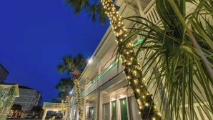 A hotel with a row of palm trees wrapped in string lights, a covered outdoor seating area, and a building with a balcony. - Sunny Studio Near the Beach at Palm Court (#6) (North Myrtle Beach)