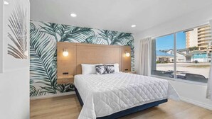 A modern bedroom with a large bed, wooden headboard, and a view of buildings through a large window. - Sunny Studio Near the Beach at Palm Court (#6) (North Myrtle Beach)