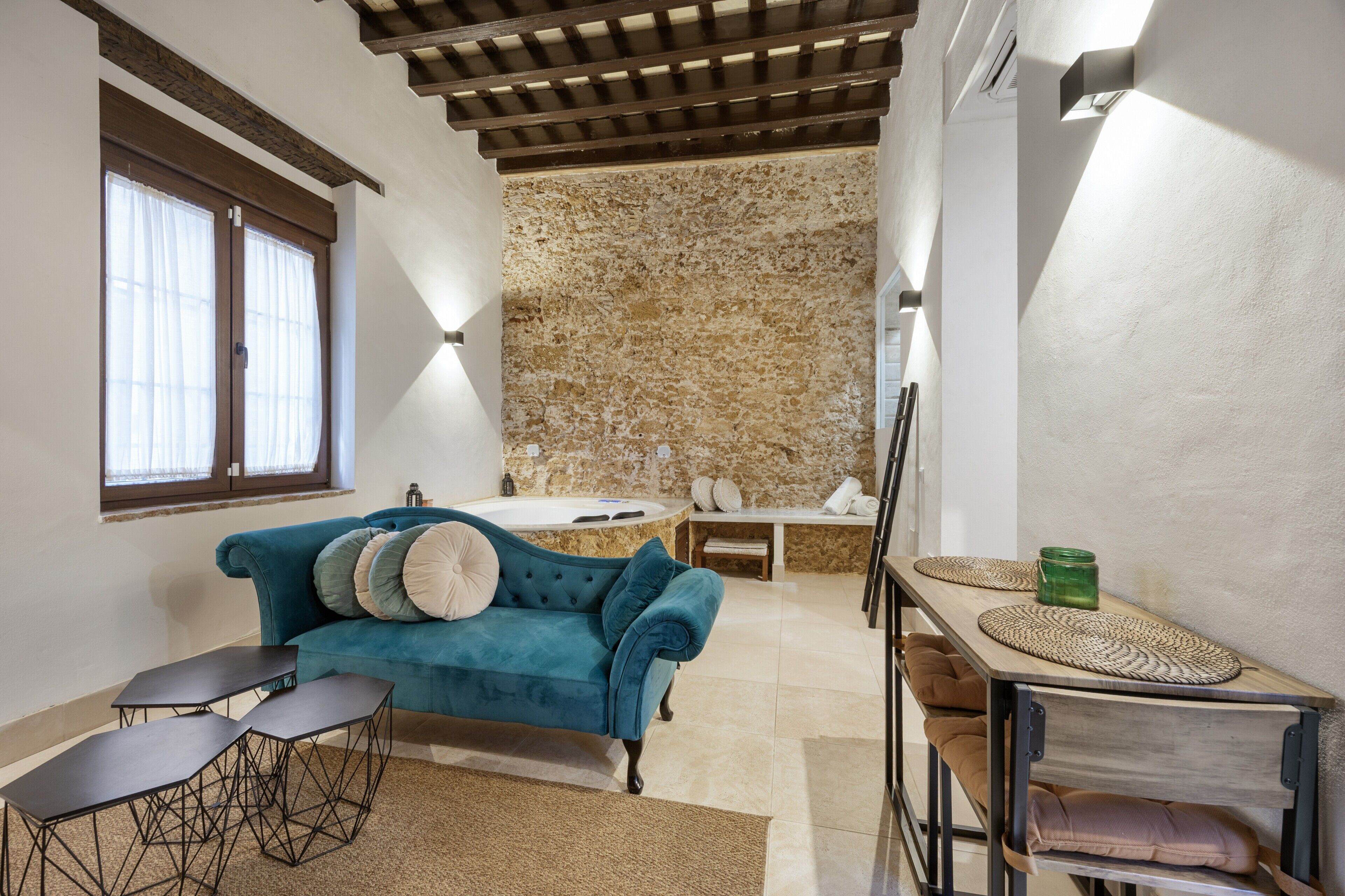 A room with a blue sofa, a small table, a bench, a wooden table with woven placemats, a stone wall, and a bathtub.