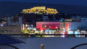 A table set for two with wine glasses and a bottle, overlooking a cityscape with a brightly lit building in the background. - Acropolis Magical View Suite, Metro Kerameikos (Athens)