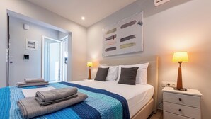A neatly made bed with a blue and white quilt, two bedside lamps, a nightstand with drawers, and a painting on the wall. - Acropolis Magical View Suite, Metro Kerameikos (Athens)