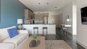 Un salon moderne comprenant un canapé, une table basse avec une plante, et un coin cuisine avec un bar et des sièges.
