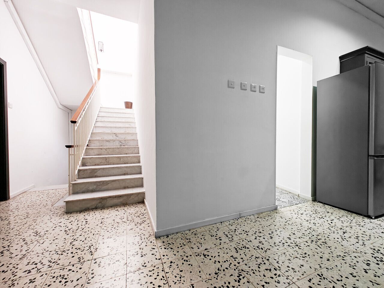 A modern kitchen with a stainless steel refrigerator, a staircase leading to a bright area, and a speckled floor.