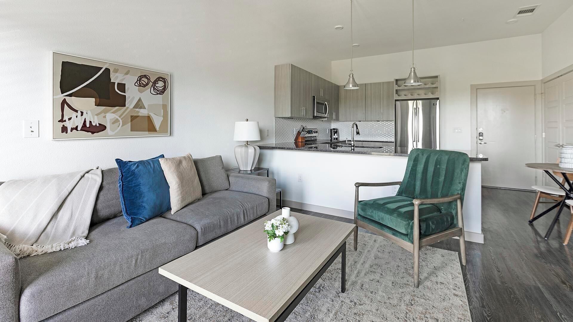 En moderne stue med en grå sofa, en grøn lænestol og et sofabord. Et køkken kan ses i baggrunden med rustfrie hvidevarer.