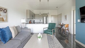 A modern living room with a grey sofa, a green armchair, and a coffee table. A kitchen is visible in the background with stainless steel appliances. - Landing | Amazing 1BD, Pool, Gym (Allen)