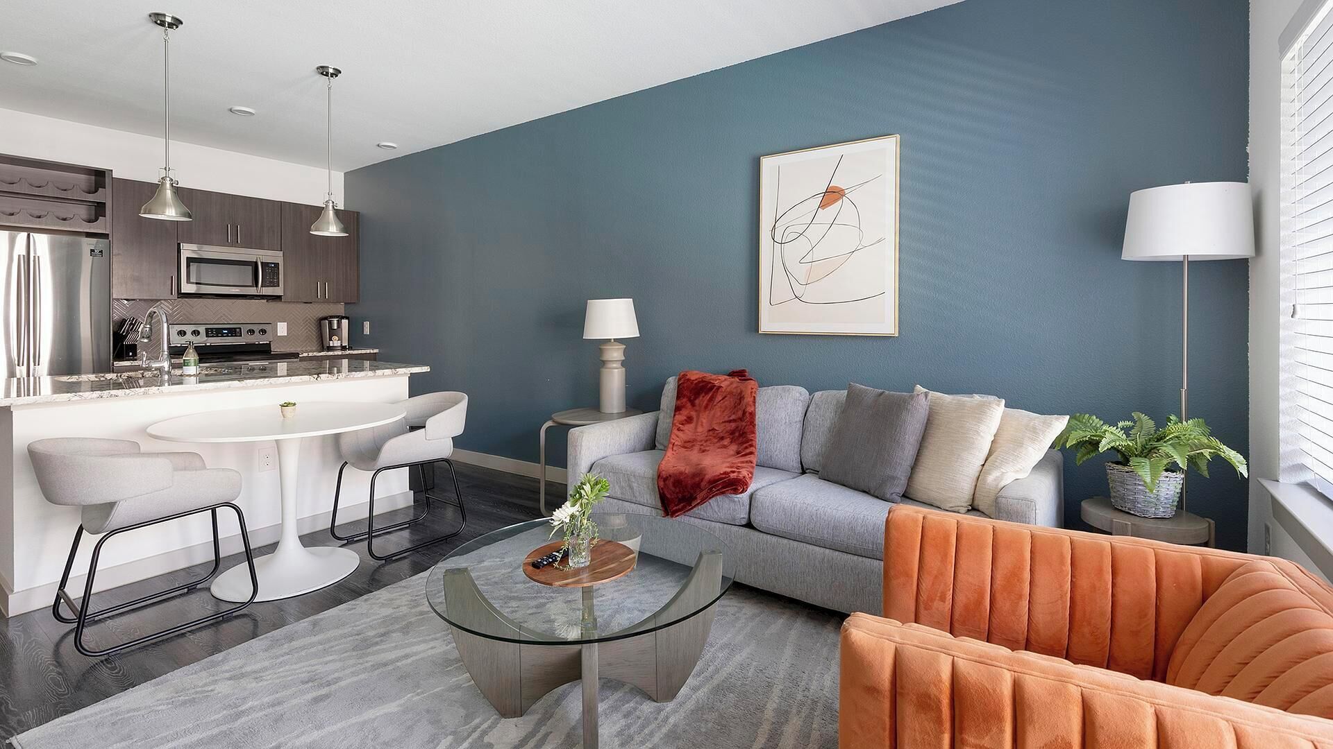 Een moderne woonkamer met een grijze bank, een oranje fauteuil, een glazen salontafel, en een keuken met een wit eiland en hanglampen.