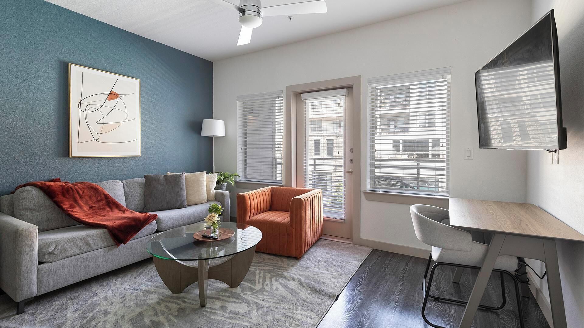 A modern living room with a grey sofa, a glass coffee table, a red armchair, and a desk with a chair. There is a ceiling fan, a floor lamp, and a large window with blinds.
