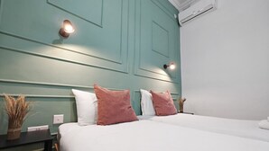 Et hotelværelse med en turkis farvet sengegavl, to senge med sengetøj i hvidt og lyserød, et natbord med en vase med tørrede planter, og en aircondition-enhed på væggen.