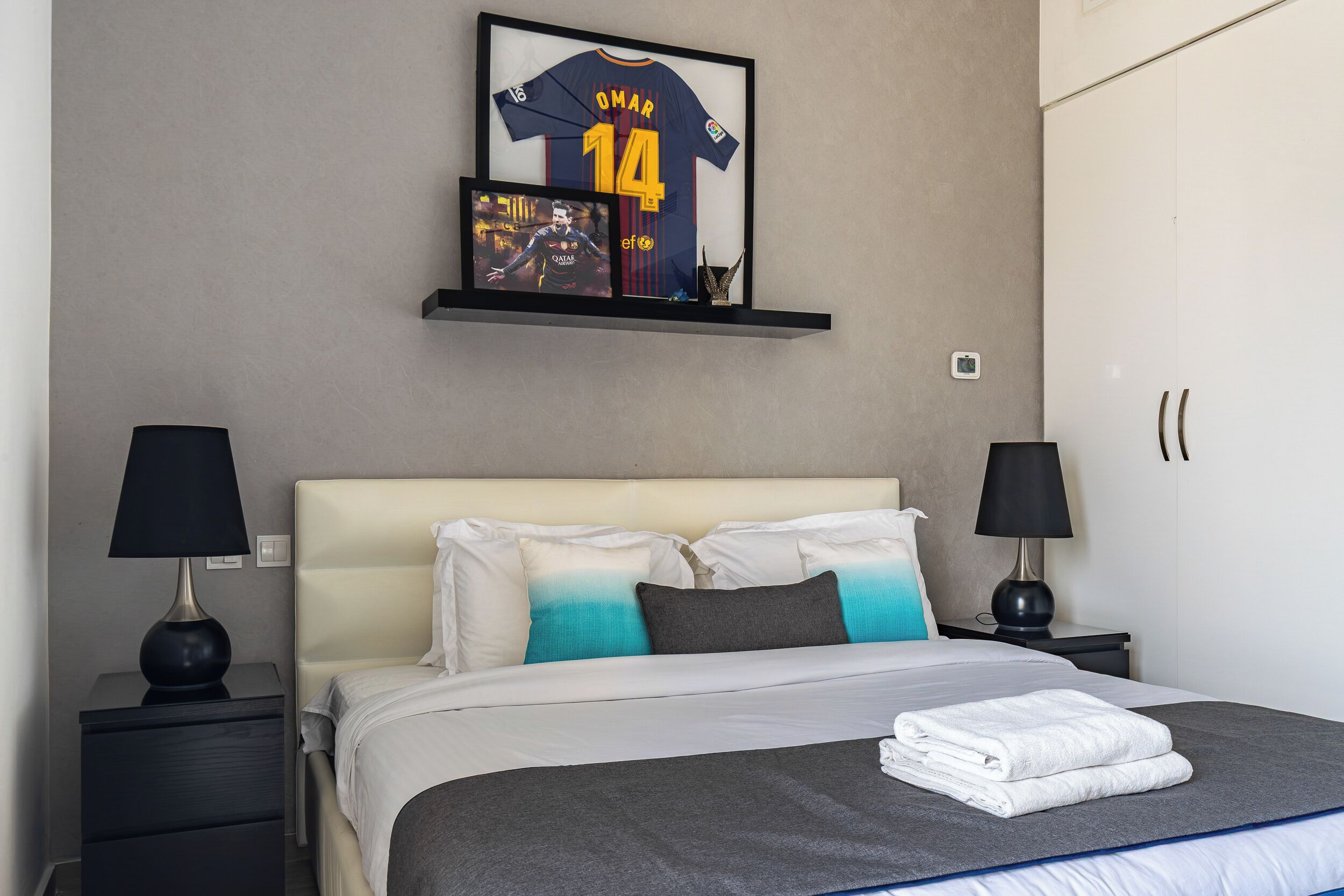 A neatly made bed with a folded towel, a shelf with a framed jersey, and two bedside tables with lamps.