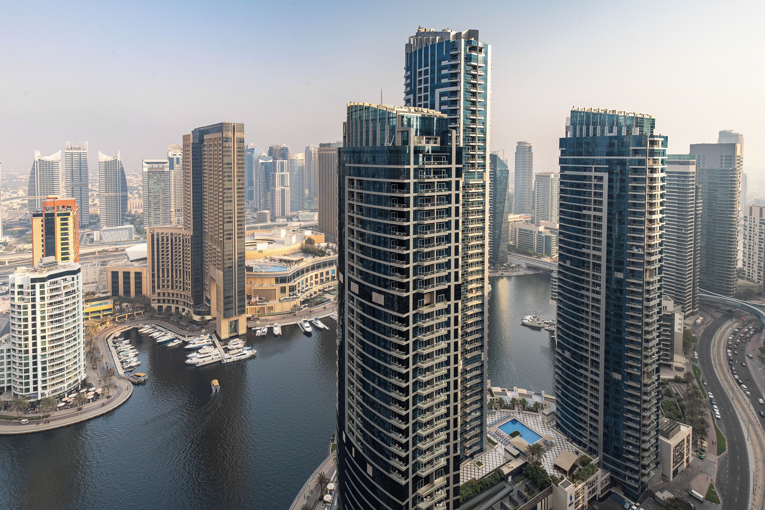 Um edifício alto com design moderno, cercado por outros edifícios altos e uma marina com barcos.