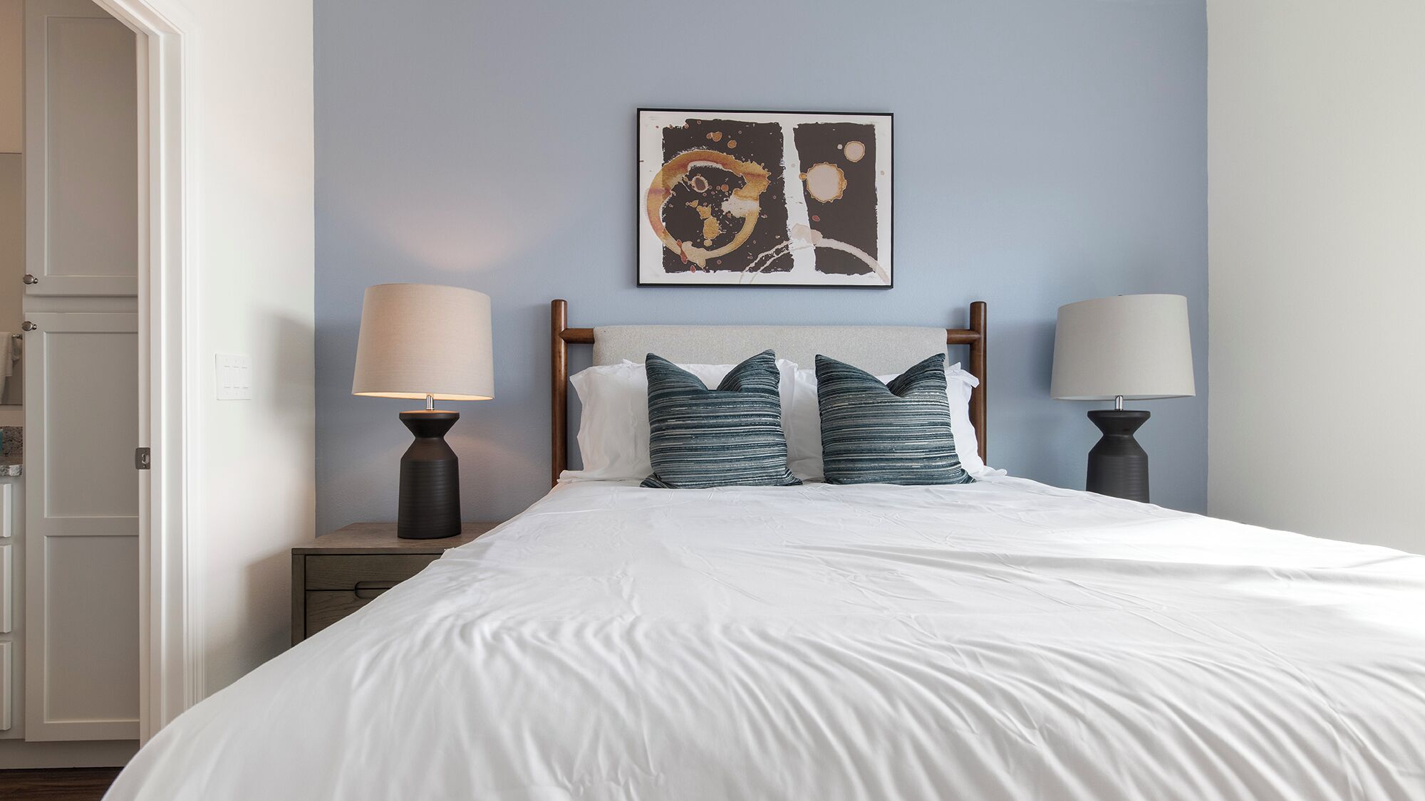 A neatly made bed with white linens, two bedside lamps, and a wall-mounted abstract artwork.