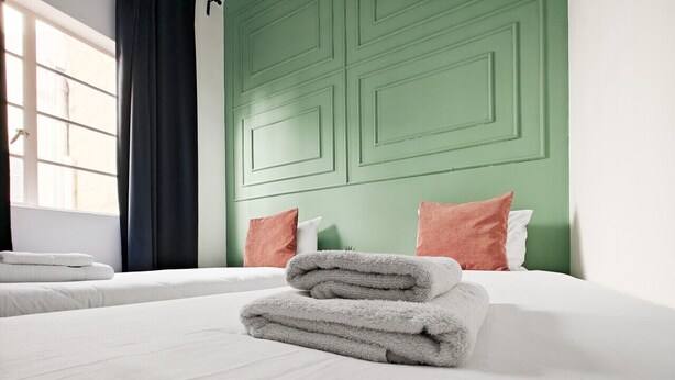 A neatly made bed with folded towels and pillows, a green accent wall, and a window with curtains. - Cosy room 7  1 bedroom (Pietà)