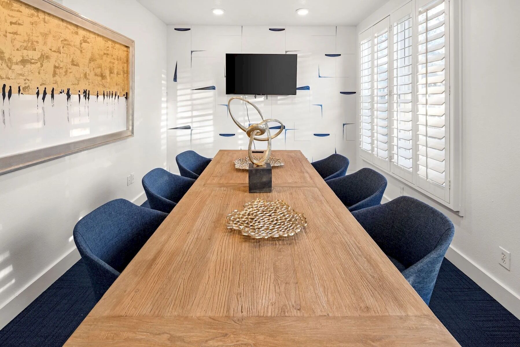 A conference room with a long wooden table, blue chairs, a mounted TV, and a wall art piece.
