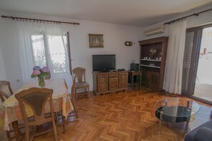 Ein Wohnzimmer mit Holzdielen im Fischgrätmuster, einem Esstisch mit Stühlen, einem Fernseher, einem Schrank und einem Glastisch.