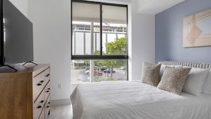 Ein Schlafzimmer mit großem Fenster, einem Bett mit weißer Bettwäsche, einer hölzernen Kommode und einem Bild an der Wand.