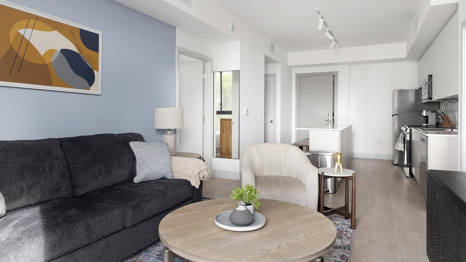 Een moderne woonkamer met een donkergrijze bank, een ronde salontafel met een plant, een lichtkleurige fauteuil en een keuken met een fornuis en een koelkast.