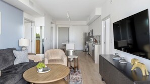 Ein modernes Wohnzimmer mit einer Couch, einem Couchtisch und einem Fernseher. Es gibt eine Küchenzeile mit Kühlschrank und Mikrowelle.
