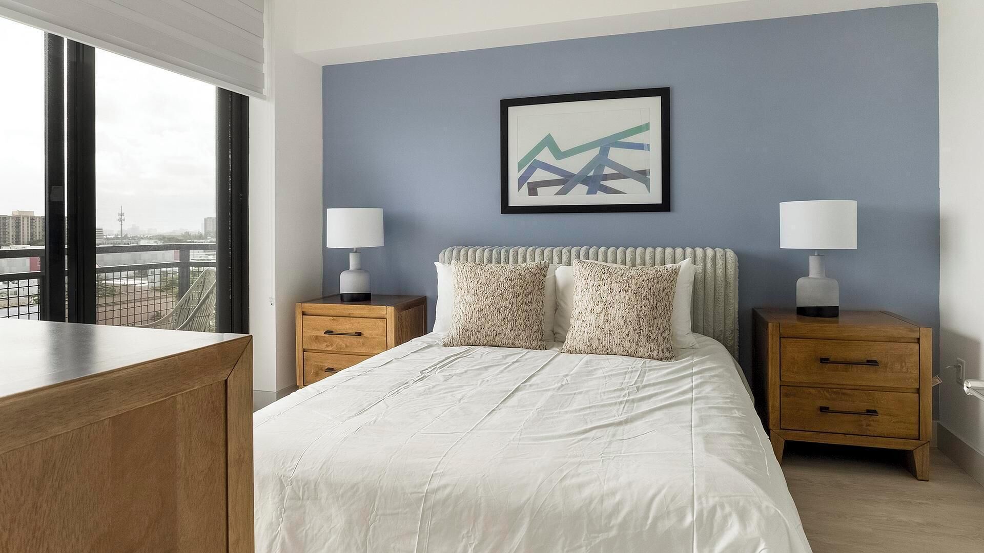 A hotel room with a bed, two bedside tables, a window with a view of the city, and a framed abstract artwork on the wall.