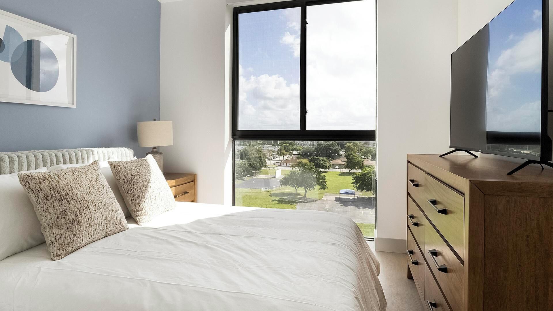 A bedroom with a large window, a bed with white bedding, a wooden dresser, and a framed picture on the wall.