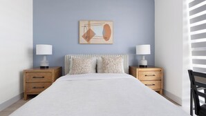 Een slaapkamer met een bed, twee nachtkastjes met lampen, een abstract schilderij aan de muur en een raam met zonwering.
