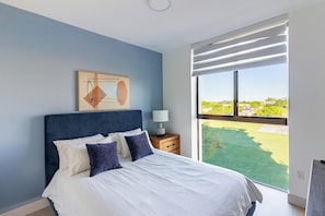 A bedroom with a large bed, a nightstand, a lamp, and a view of a green landscape through a window. - Landing | Stunning 2BD, Yoga Studio, Clubhouse (North Miami)