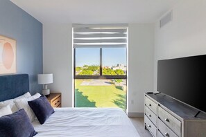 A bedroom with a bed, a TV, a nightstand, a lamp, and a view of a green lawn and buildings. - Landing | Beautiful 2BD, Clubhouse, Yoga Studio (North Miami)